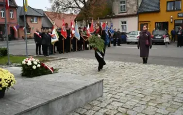 widzimy 2 panie idące z wieńcem w strone pomnika, w tle widoczne są sztandary