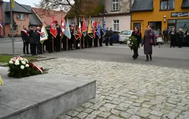widzimy 2 panie idące z wieńcem w strone pomnika, w tle widoczne są sztandary