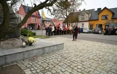 widzimy pana Burmistrza trzymającego w ręce wieniec oraz grupe os&oacute;b stojącą ze sztandarami