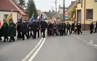 widzimy idącą po ulicy orkiestre, sztandary oraz grupe os&oacute;b