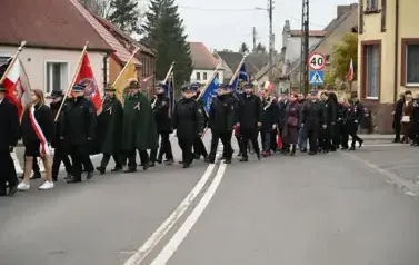 widzimy idącą po ulicy orkiestre, sztandary oraz grupe os&oacute;b
