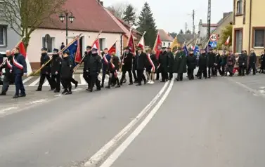 widzimy idącą po ulicy orkiestre, sztandary oraz grupe os&oacute;b