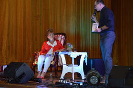 Na zdjęciu widać panią Czesię siedzącą na scenie na bardzo eleganckim fotelu, obok stoi prowadzący.