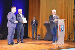 Na zdjęciu dw&oacute;ch mężczyzn w granatowych garniturach stoi na scenie. Jeden z nich trzyma nagrodę. Trzeci mężczyzna przemawia przy m&oacute;wnicy.