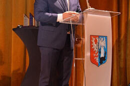 Na zdjęciu mężczyzna w ciemnym garniturze wygłasza przemowę przy m&oacute;wnicy.