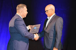 Na zdjęciu mężczyzna wręcza nagrodę mężczyźnie w okularach. Oboje są ubrani w eleganckie, granatowe garnitury. 