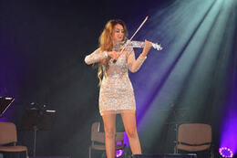 Na zdjęciu znajduje się kobieta w srebrnej sukience. Gra na szklanych skrzypcach.