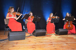 Na zdjęciu widać cztery kobiety w długich czerwonych sukniach. Siedzą na krzesłach i grają na skrzypcach.