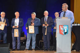 Na zdjęciu widać przemawiającego mężczyznę przy m&oacute;wnicy. Za nim stoją inni eleganccy panowie trzymający w rękach nagrody.