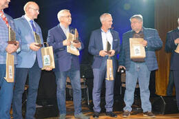 Na zdjęciu widać pięciu meżczyzn na scenie. W rękach trzymają statuetki i nagrody. Ubrani są elegancko.