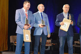 Na zdjęciu widać trzech mężczyzn na scenie. W rękach trzymają nagrody. Ubrani są elegancko.