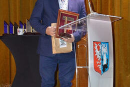 Na zdjęciu znajduje się mężczyzna w granatowym garniturze. Stoi przy m&oacute;wnicy. W ręce trzyma nagrody.