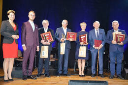 Na zdjęciu od lewej strony znajduje się przewodnicząca Rady Gminy ubrana w czerwoną sukienkę i czarny żakiet, obok niej stoi w&oacute;jt gminy, następnie pięciu nagrodzonych przedsiębiorc&oacute;w. W tym jedna kobieta. Wszyscy ubrani w granatowe garnitury, a kobieta w 