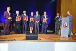 Na zdjęciu widać sześć os&oacute;b stojących na scenie. Od lewej strony w&oacute;jt gminy, następnie czterech meżczyzn i jedna kobieta. Wszyscy trzymają nagrody i odznaczenia Herbu Powiatu Wągrowieckiego. Kobieta ubrana jest w granatową ssukienkę, mężczyżni są w granat