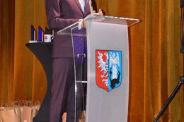 Na zdjęciu widać przemawiającego na scenie w&oacute;jta. Stoi przy m&oacute;wnicy. Ubrany jest w śliwkowy garnitur.