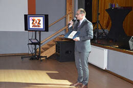 Na zdjęciu widać mężczyznę w okularachw  garniturze, z mikrofonem w ręce. Stoi przed sceną i prowadzi imprezę.