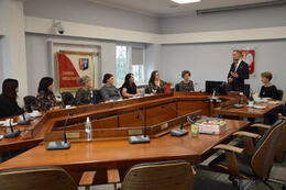 Na zdjęciu widać osiem kobiet siedzących wok&oacute;ł stołu. Pan w&oacute;jt stojąc przemawia.