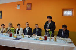 Zdjęcie przedstawia st&oacute;l a za nim siedzących gości. Kobieta stoi i wygłasza przemowę. Ubrana jest w wzorzystą sukienkę i ciemną marynarkę.