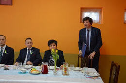 Zdjęcie przedstawia st&oacute;l a za nim siedzących gości. Jeden mężczyzna stoi i wygłasza przemowę. Ubrany jest w granatowy garnitur. W ręce trzyma granatową teczkę.