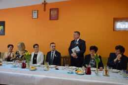 Zdjęcie przedstawia st&oacute;l a aza nim siedzących gości. Jeden mężczyzna stoi i wygłasza przemowę. Ubrany jest w granatowy garnitur. W ręce trzyma książkę.