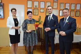 Zdjęcie przedstawia od lewej strony przeowdniczącą Rady Gminy ubraną w czarną sukienkę i biały żakiet. Obok stoi pani Sołtys obchodząca jubileusz, trzyma w ręce grawerton i kwiaty. Następnie jest w&oacute;jt Gminy Mieścisko w granatowym garniturze. 