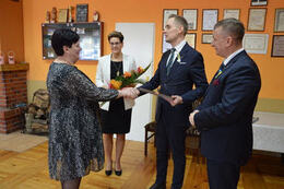 W&oacute;jt wręcza grawerton pani sołtys kt&oacute;ra obchodzi jubileusz 20lecia bycia sołtysem. Kobieta ubrana jest w ciemną sukienkę, w&oacute;jt w granatowy garnitur. Obok stoją zaproszeni goście.