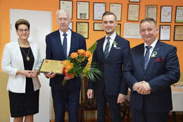 Zdjęcie przedstawia od lewej strony przeowdniczącą Rady Gminy ubraną w czarną sukienkę i biały żakiet. Obok stoi Sołtys obchodzący jubileusz, trzyma w ręce grawerton i kwiaty. Następnie jest w&oacute;jt Gminy Mieścisko w granatowym garniturze. Z prawej strony st