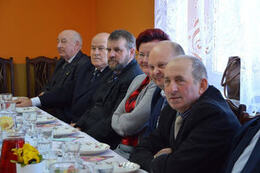 Zdjęcie przedstawia ludzi siedzących przy stole. Mężczyźni ubrani w ciemne marynarki. Po środku siedzi kobieta w czerwonej koszuli i swetrze.