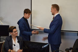 Na zdjęciu w&oacute;jt wręcza dokumenty i gratulacje młodemu radnemu. Jest to chłopak w kr&oacute;tkich włosach. Ubrany w ciemną koszulę i granatową marynarkę.