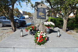 Zdjęcie przedstawia pomnik ofiar II Wojny Światowej. Leży na nim wiązanka kwiat&oacute;w, w wazonie znajduje się białoczerwony bukiet, stoją dwa znicze.