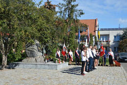 Zdjęcie przedstawia pomnik upamiętniający ofiary II Wojny Światowej. Przed nim stoi kilkoro uczni&oacute;w prezentujących program artystyczny. W tle widać delegacje ze sztandarami.