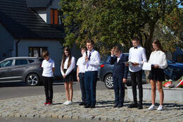 Zdjęcie przedstawia młodzież w eleganckich ubraniach. Jeden z chłopak&oacute;w trzyma w ręce mikrofon i recytuje wiersz.