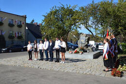 Na zdjęciu widać siedmioro uczni&oacute;w stojących przed pomnikiem. Prezentują przygotowany występ artystyczny. Wszyscy ubrani są na galowo.