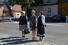 Zdjęcie przedstawia trzy kobiety stojące przed pomnikiem. Są one elegancko ubrane. Dwie mają suknie w kwiaty, trzecia granatową i biały żakiet.