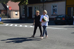 Zdjęcie przedstawia delegację jednostek pomocniczych gminy niosącą bukeit kwiat&oacute;w. Dwie kobiety idą obok siebie. Jedna ubrana na czarno, druga w jasną koszulę i ciemne spodnie.