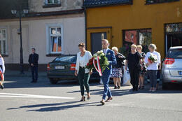 Zdjęcie przedstawia w&oacute;jta Gminy Mieścisko wraz z przewodniczącą Rady Gminy niosoących wiązankę. Mężczyzna ubrany w granatowy garnitur, kobieta białą bluzkę i jasny żakiet oraz zielone spodnie.