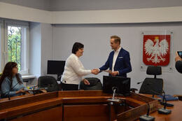 Zdjęcie przedstawia w&oacute;jta gminy podającego dłoń opiekuńce ukraińskiej młodzieży. Kobieta ma kr&oacute;tkie, ciemne włosy, jasny sweter. W&oacute;jt ma granatową marynarkę. Oboje są uśmiechnięci.