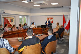 Na zdjęciu widać st&oacute;ł na sali sesyjnej przy kt&oacute;rym siedzą ludzie. Są to zar&oacute;wno dorośli jak i młodzież. Po środku stoi w&oacute;jt i przemawia.