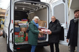 Na zdjęciu widać białego busa zapakowanego prezentami. Starsza kobieta i mężczyzna wyjmują z niego preznety.