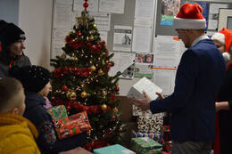 Zdjęcie przedstawia W&oacute;jta w czapce Mikołaja. Trzyma w ręce prezent. Obok stoją dzieci w kurtkach i czapkach.