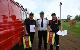 Strażacy stoją obok wozy strażackiego trzymając zaświadczenie o ukończeniu kursu oraz plecak z wyposażeniem medycznym