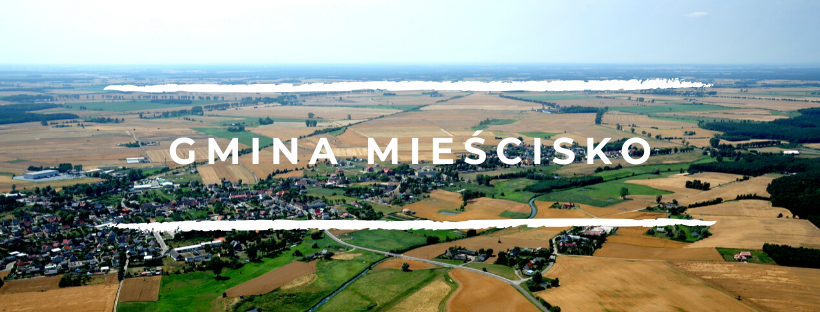 Na zdjęciu widac panoramę Mieściska - pola, małe domki w oddali. Na pierwszym planie jest duży biały napis "Gmina Mieścisko"
