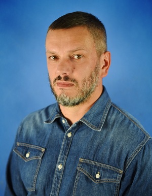 Zdjęcie przedstawia portret Radnego Tomasza Kozłowskiego. Mężczyzna ma krótkie włosy, siwy zarost. Ubrany jest w jeansową koszulę.